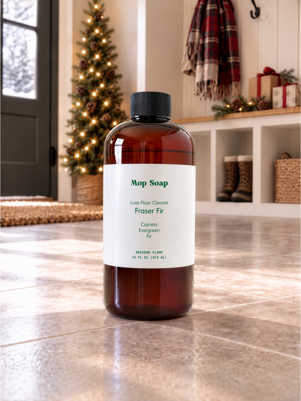 A 16oz amber bottle of Mop Soap 'Fraser Fir' luxury floor cleaner with a white label and black cap, set on a tiled floor in a cozy, holiday themed room with a Christmas tree in the background.