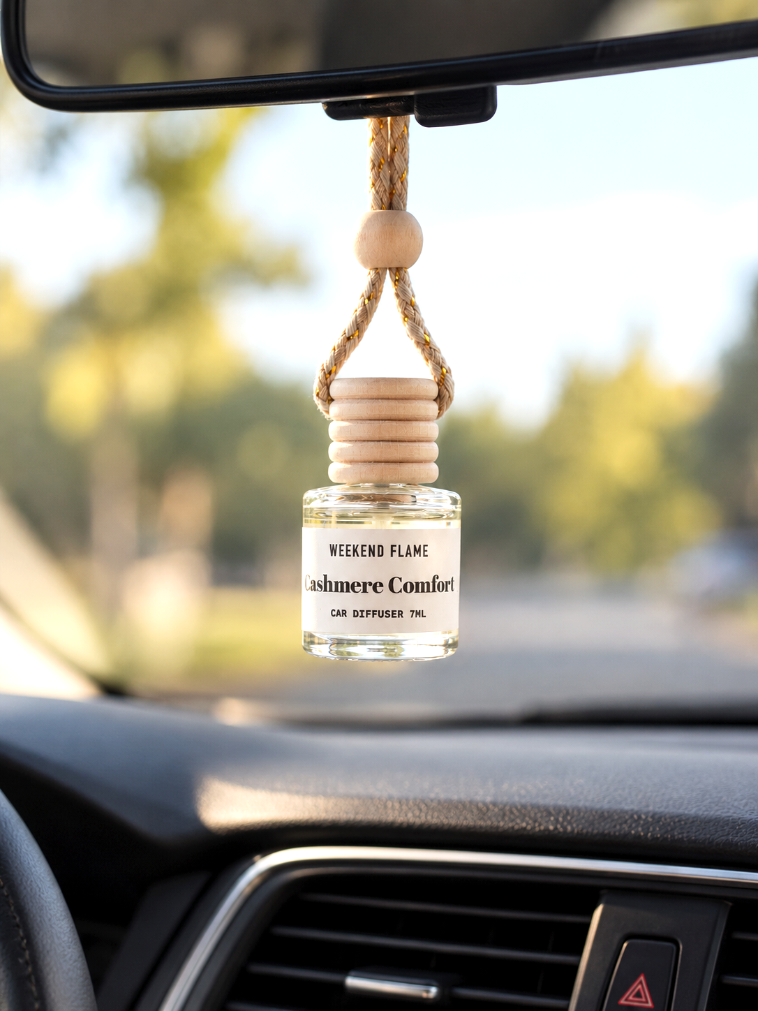 Scented hanging car diffuser hangs from a rearview mirror inside a car. The clear glass bottle contains fragrance oil and has a white label that reads 'WEEKEND FLAME' and 'Cashmere Comfort Car Diffuser 7ml.