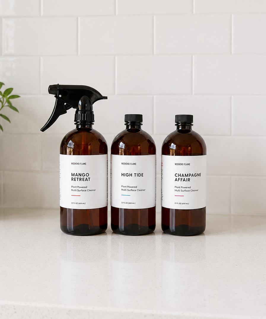 Three amber plastic bottles of luxury all purpose cleaning spray on a white countertop against a white subway tile backsplash. The bottles have white labels with black text and are topped with black caps, including a spray bottle on the left and two closed bottles on the right.