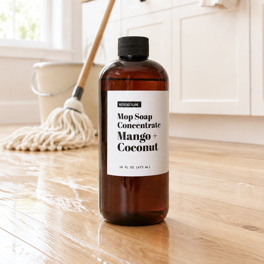A 16 oz amber bottle of Mop Soap Concentrate in Mango and Coconut scent sitting on a clean, shiny hardwood floor with a mop in the background.