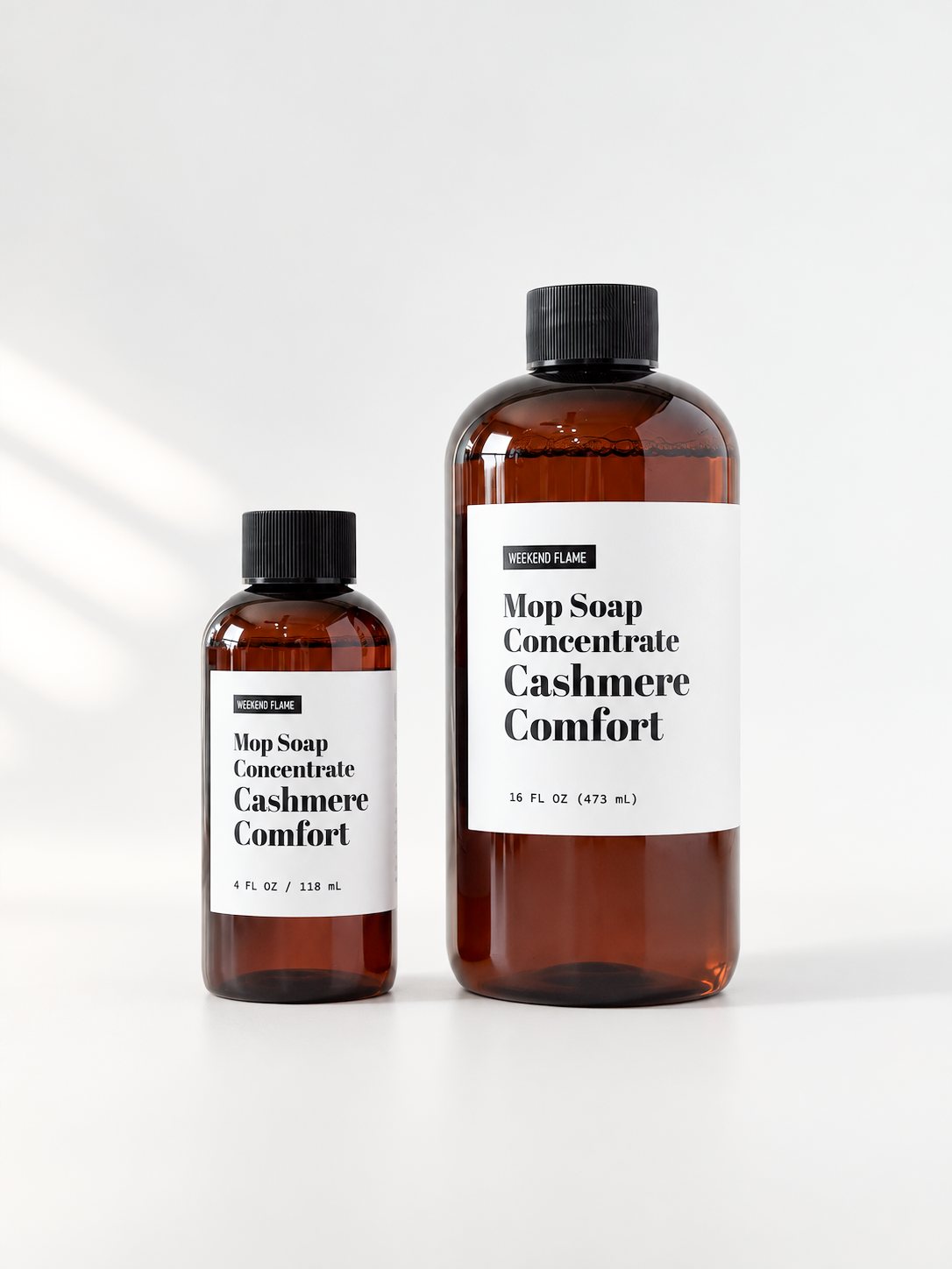 Two amber plastic bottles of Mop Soap Concentrate in the Cashmere Comfort scent, one 4 fl oz and one 16 fl oz, with black caps and white labels, on a clean white background.