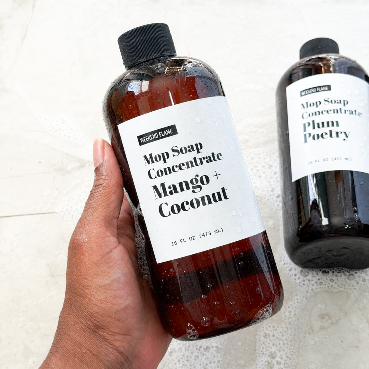 A hand holds a amber bottle of Mop Soap Concentrate Mango + Coconut, with a second bottle labeled Plum Poetry in the background on a foamy light surface.