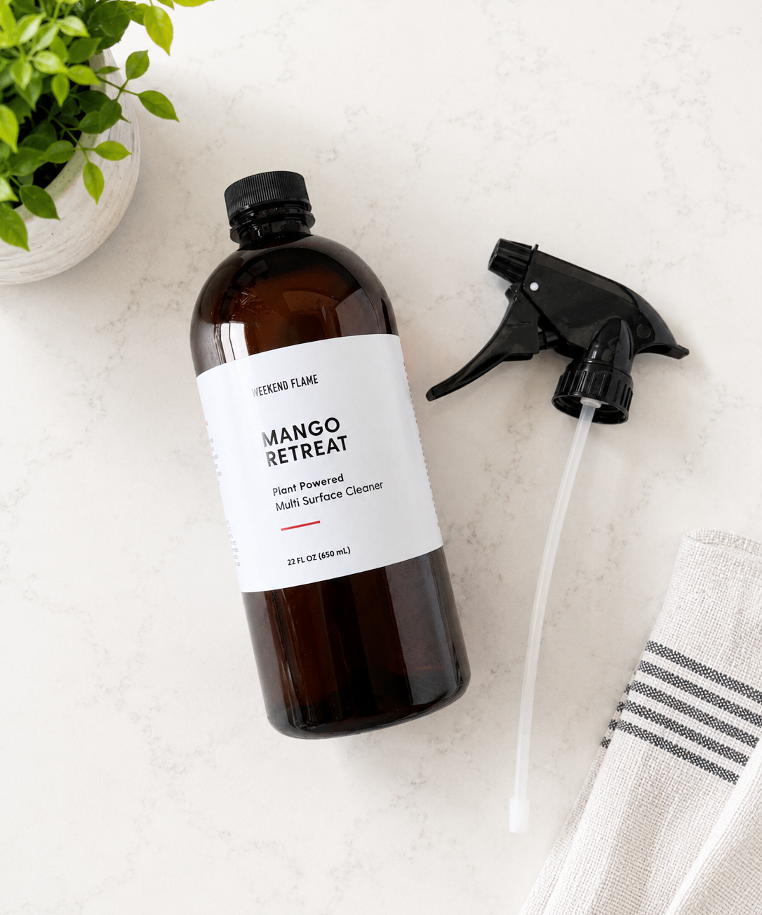 A 22 fl oz amber PET bottle of Plant Based Multi Surface Cleaner, shown with its black spray nozzle on a light marble countertop next to a green plant and a striped towel.