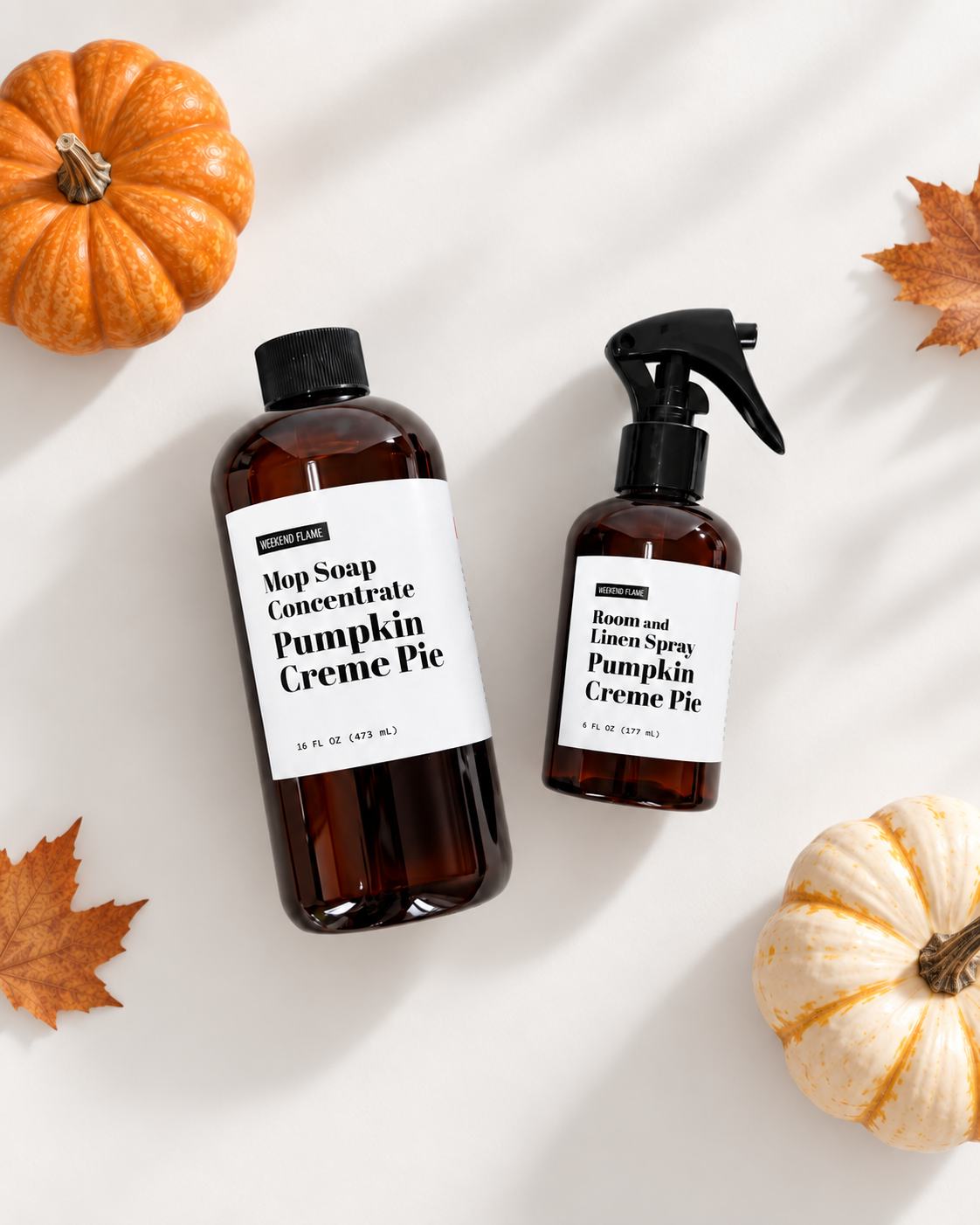 Two amber home fragrance bottles with black caps and white labels reading 'Pumpkin Creme Pie' are arranged on a white surface with autumn leaves and small pumpkins. One bottle is a mop soap concentrate and the other is a room and linen spray.