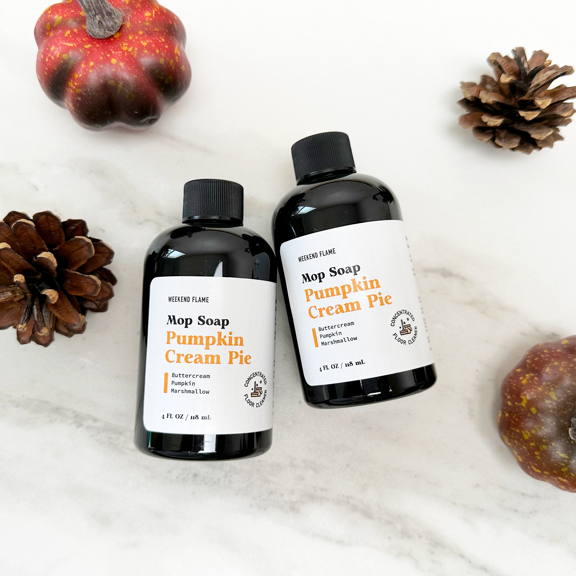 Two bottles of 'Pumpkin Cream Pie' soap on a white surface with pumpkins and pinecones.