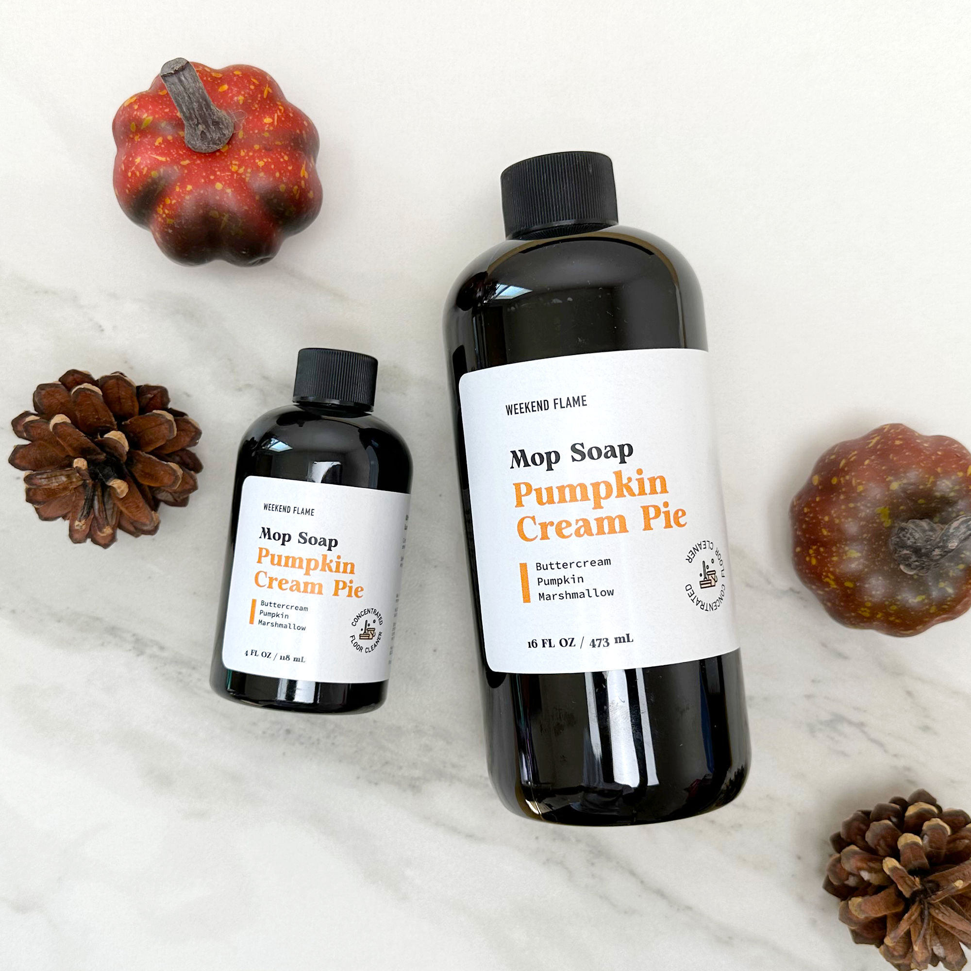 Two bottles of 'Pumpkin Cream Pie' mop soap with decorative pumpkins and pinecones on a white surface.