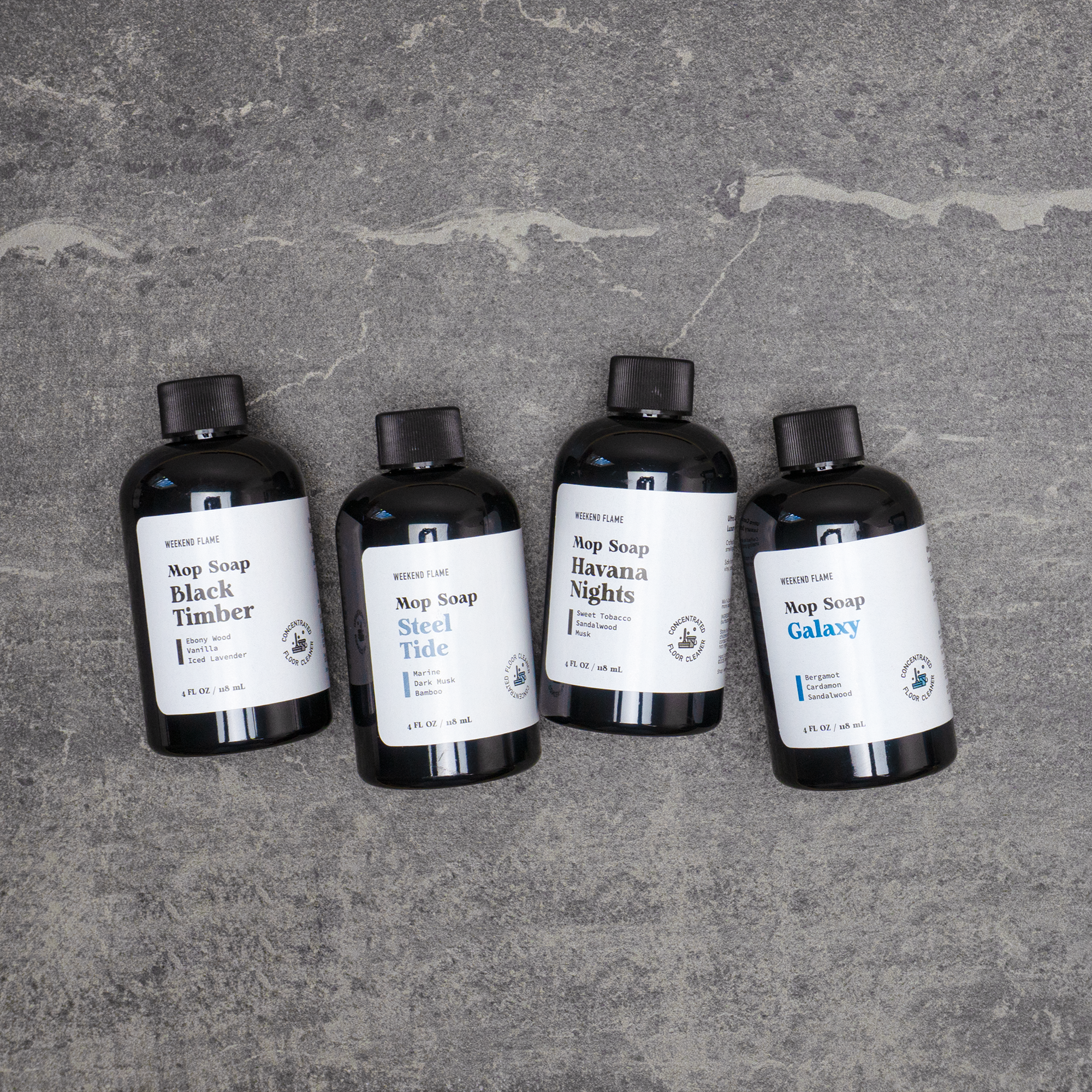 Four trial size bottles of soap labeled 'Black Timber', 'Steel Tide', 'Havana Nights', and 'Galaxy' on a gray stone surface.