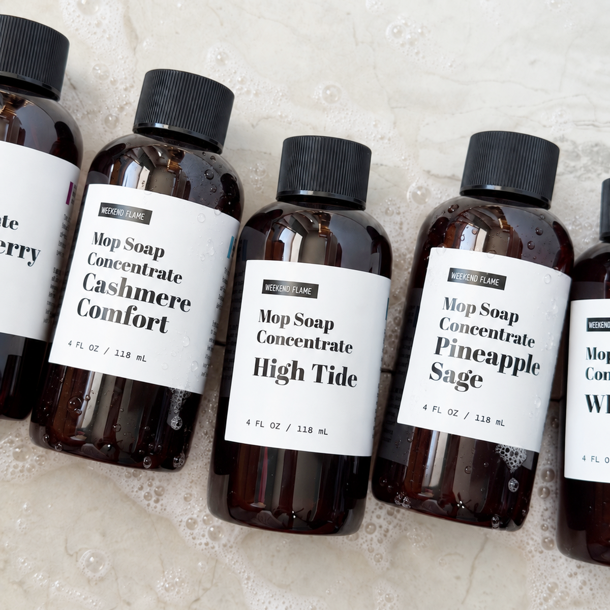 Several amber plastic bottles of Mop Soap Concentrate with black caps and white labels, arranged on a light beige textured surface with soap bubbles. Visible labels include Cashmere Comfort, High Tide, and Pineapple Sage.