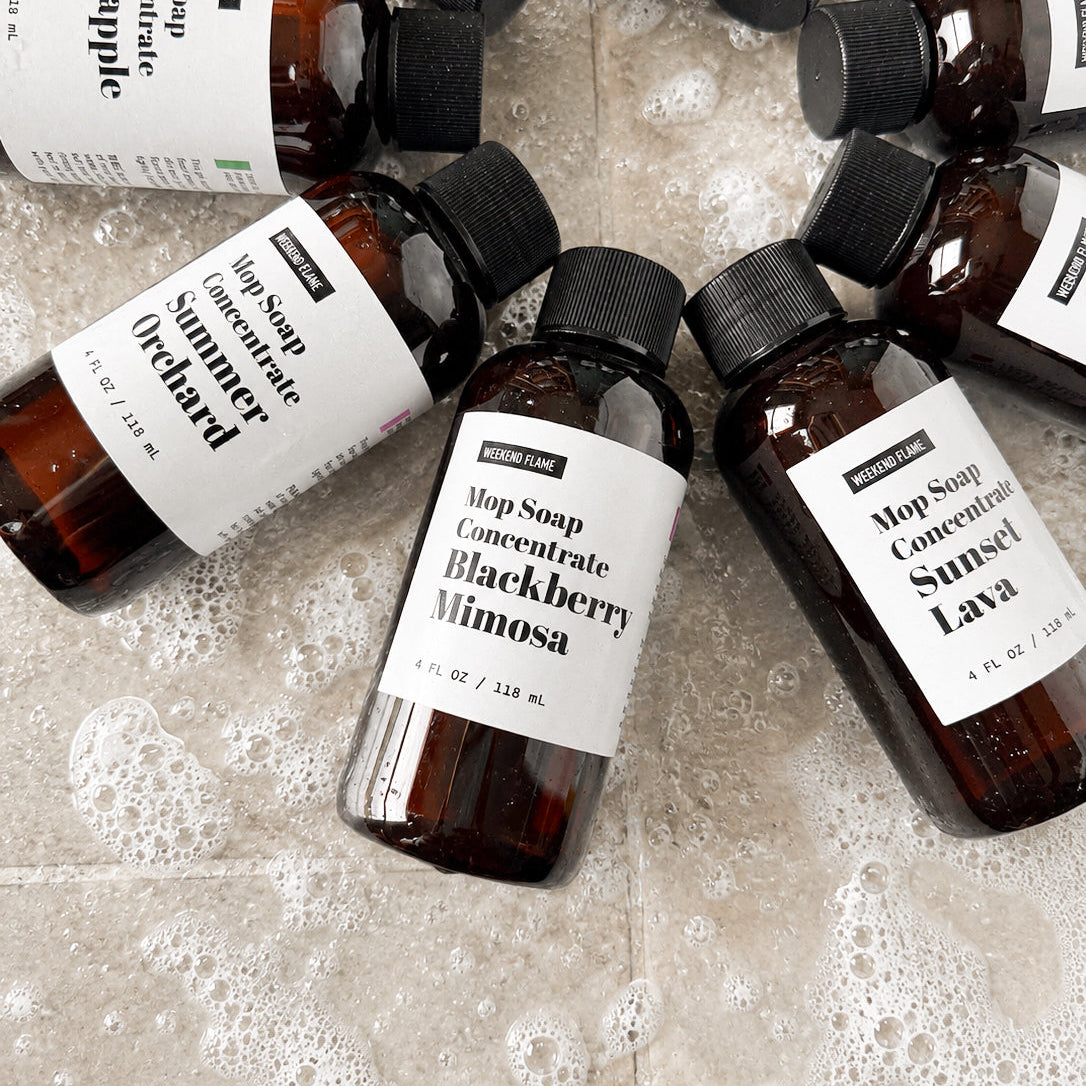 Several amber mop soap concentrate bottles with black caps and white labels are arranged in a circle on a bubbly light beige tile floor. The visible labels include Summer Orchard, Blackberry Mimosa, and Sunset Lava.