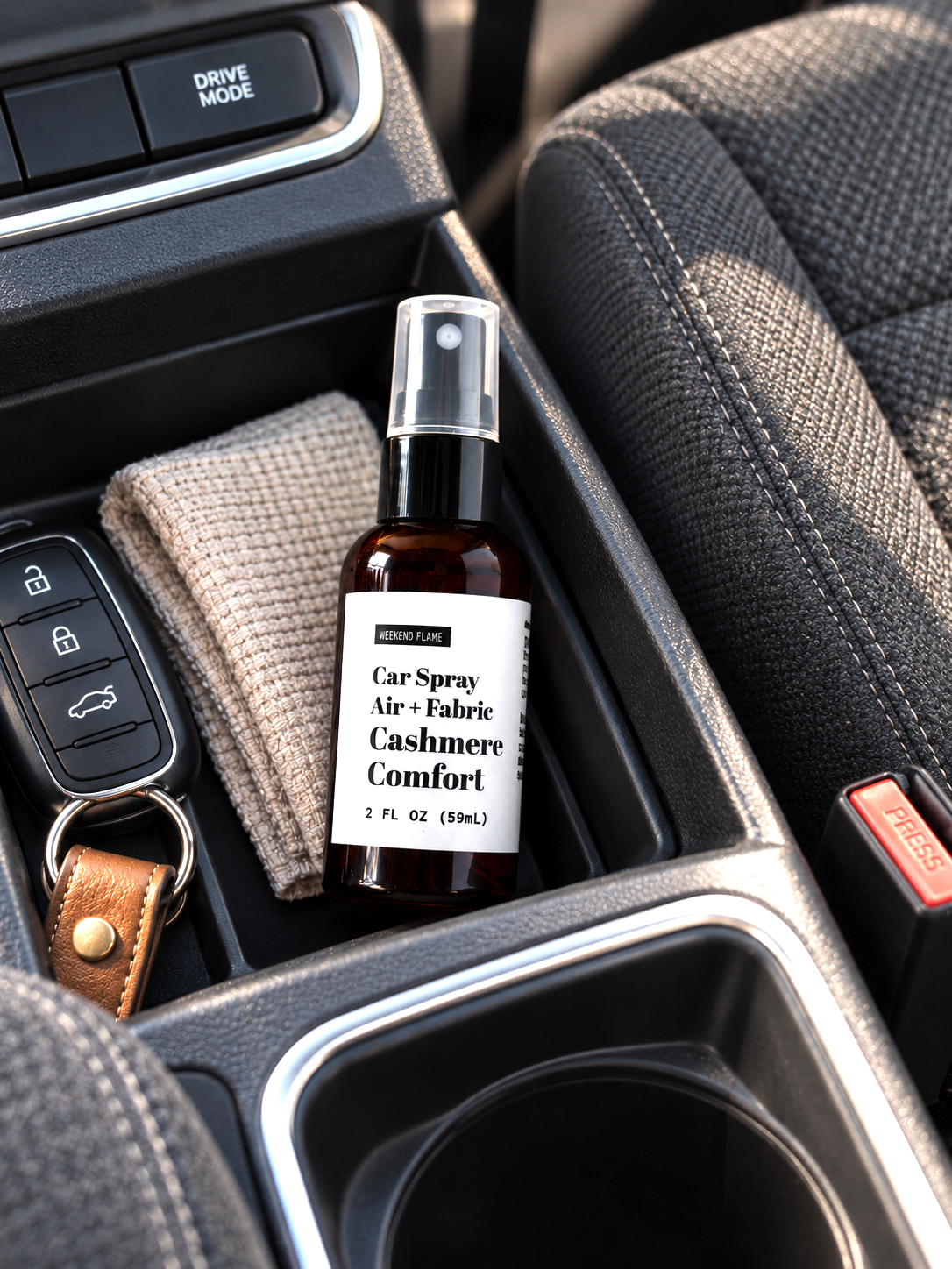 A 2 fl oz car odor eliminator spray bottle labeled 'Car Spray Air + Fabric Cashmere Comfort' resting in a car console next to a key fob and a beige cloth.