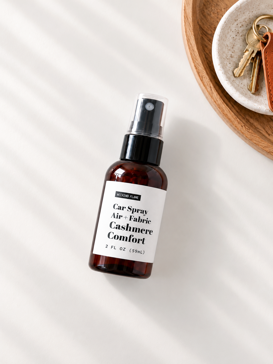 A 2 fl oz car spray bottle with a black spray top and a white label reading 'Car Spray Air + Fabric Cashmere Comfort' by Weekend Flame, styled on a white surface with soft shadows and a wooden tray with keys in the corner.