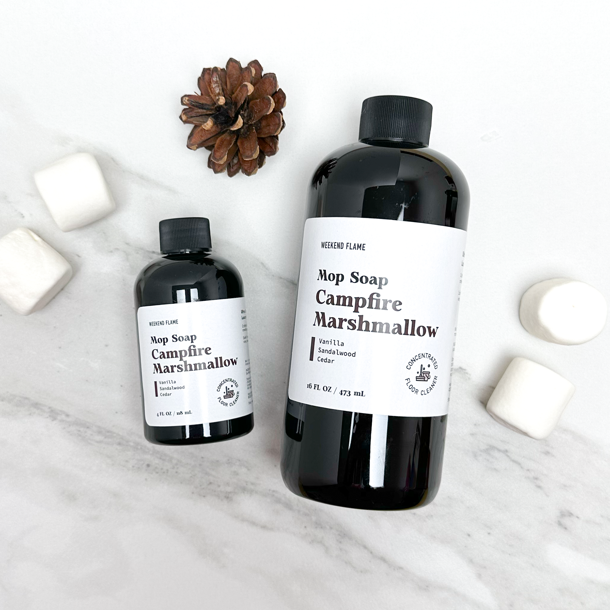 Two bottles of 'Campfire Marshmallow' fall scented mop soap on a marble surface with marshmallows and a pine cone.