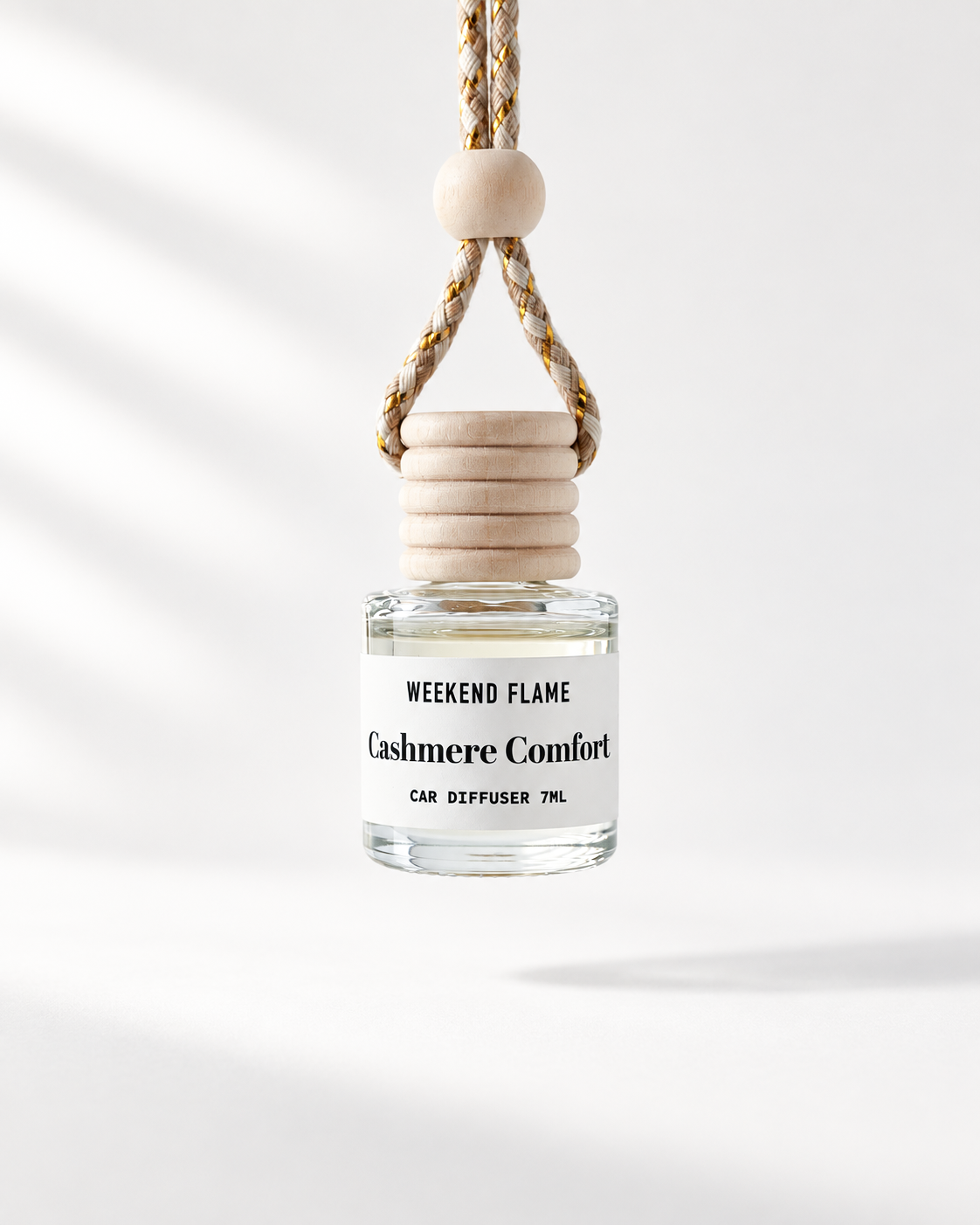A car diffuser bottle with a clear glass body, white label reading 'WEEKEND FLAME Cashmere Comfort CAR DIFFUSER 7ML', a stacked light wood cap, and a braided hanging cord with a round wooden bead, shown on a clean white background with soft shadowing.