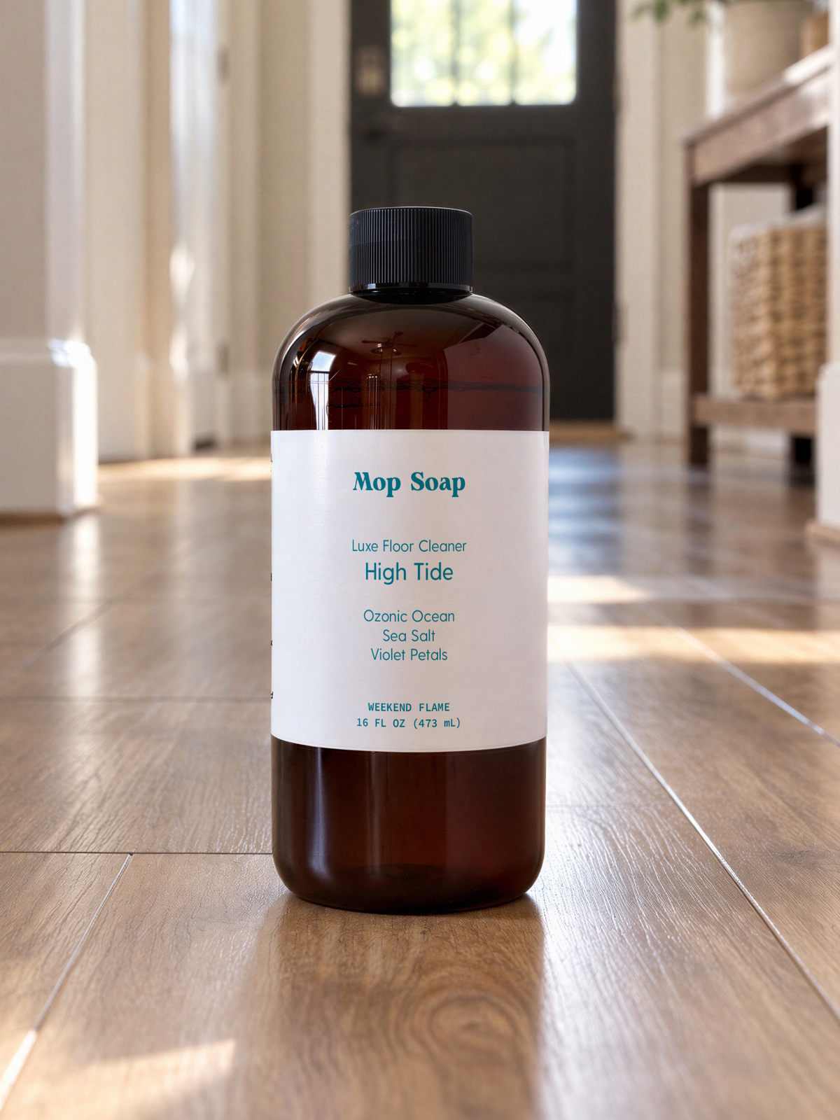 An amber bottle of Mop Soap 'High Tide' luxe floor cleaner with a white label and black cap, standing on a wooden floor in a brightly lit room.