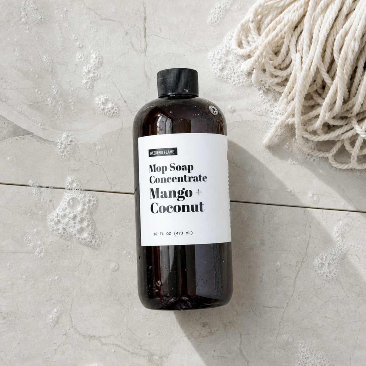 An amber glass bottle of Mop Soap Concentrate in Mango + Coconut scent, featuring a white label and black cap, styled on a light stone floor with soap bubbles and a beige rope prop.