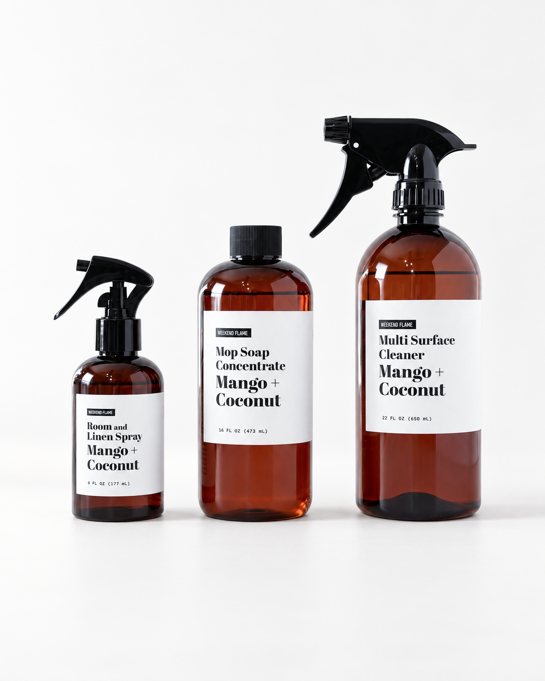 Three amber cleaning products on a white background: a small spray bottle labeled Room and Floor Spray, a medium bottle labeled Mop Soap Concentrate, and a large spray bottle labeled Multi Surface Cleaner. All have white labels and black sprayers/caps.