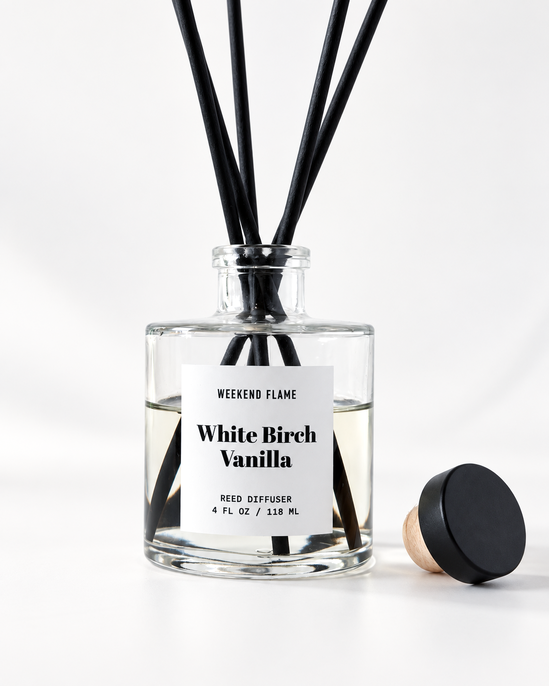 A clear glass reed diffuser bottle labeled 'Weekend Flame White Birch Vanilla' with black reeds and a separate black cap resting beside it on a clean white background.