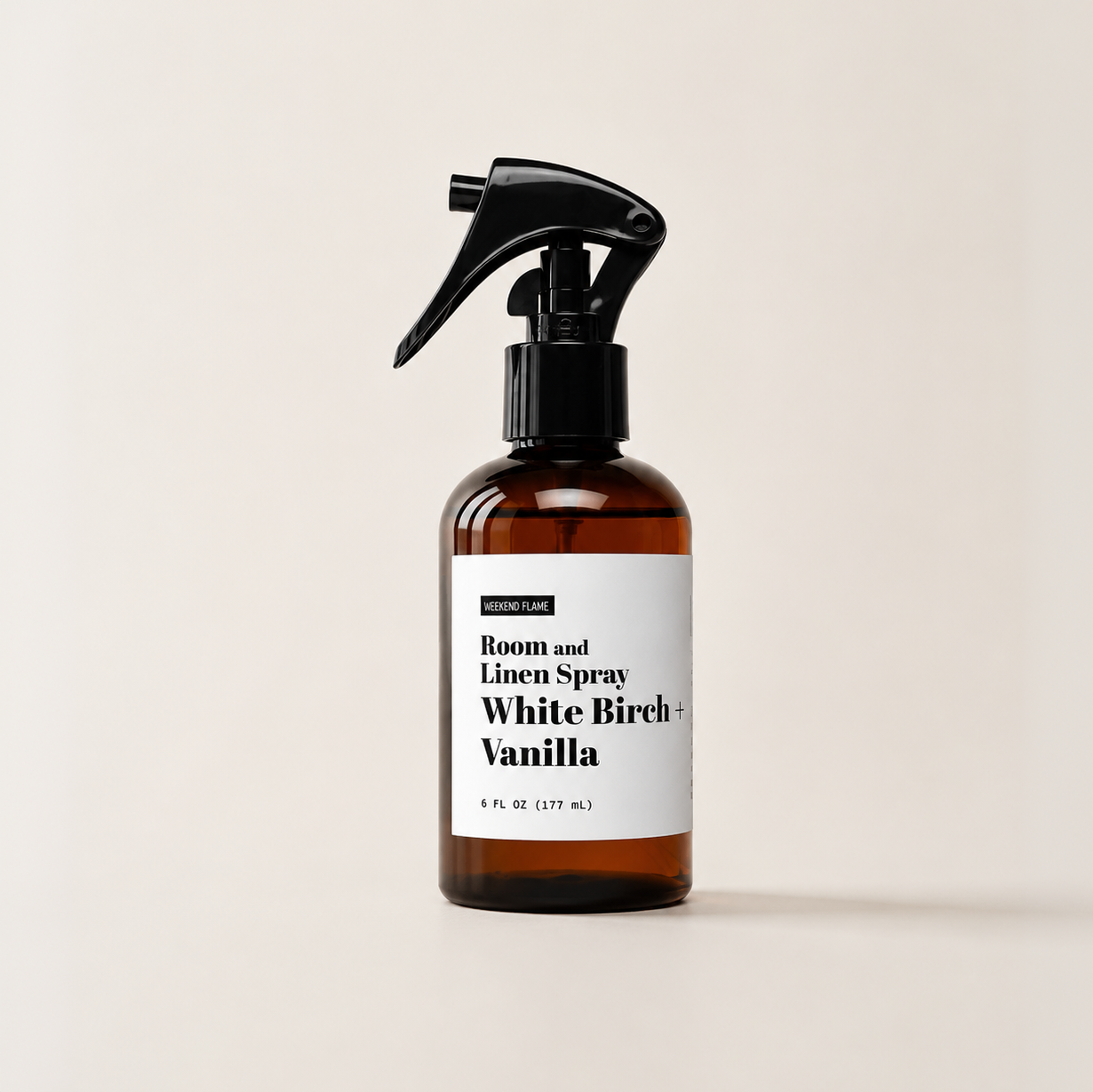 An amber plastic spray bottle with a black trigger sprayer and a white label reading 'Room and Linen Spray White Birch + Vanilla' on a warm off-white background.