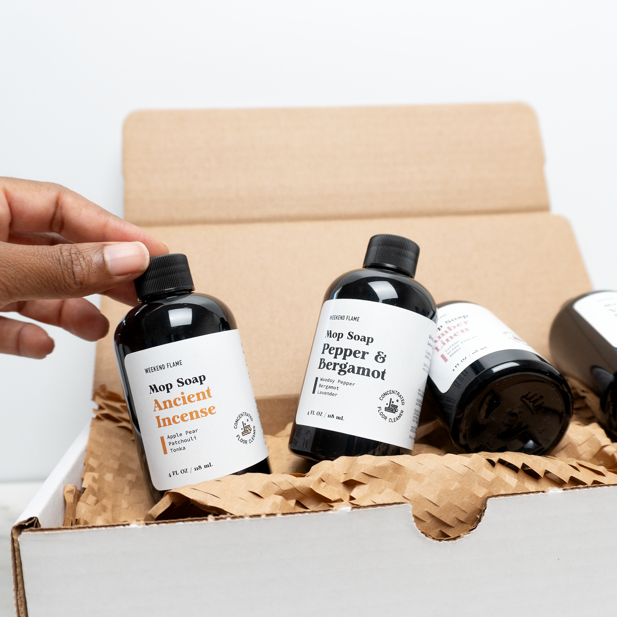 Three bottles of soap with labels in a cardboard box, one being picked up by a hand.