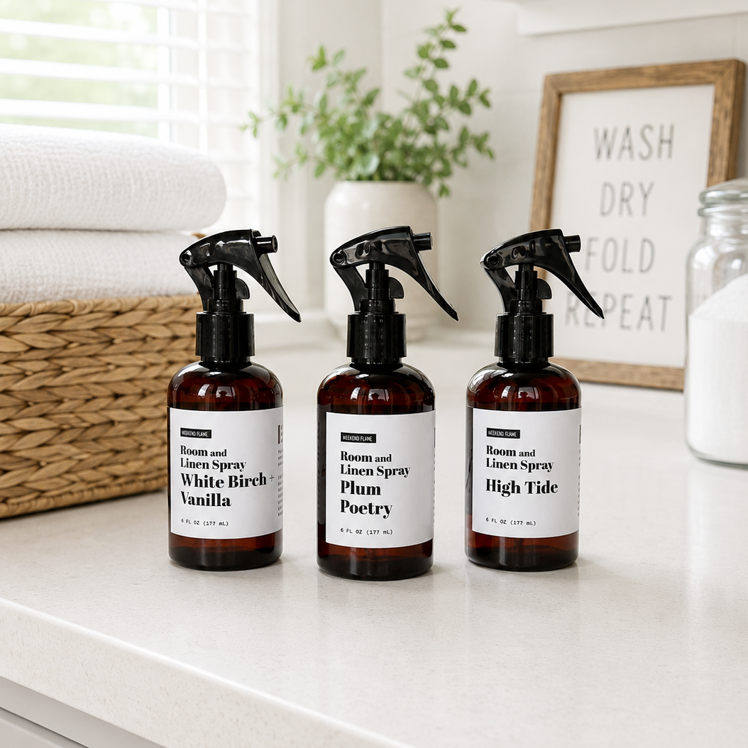 Three amber spray bottles of room and linen spray with black triggers and white labels, arranged on a white countertop in a bright laundry room setting with folded towels, a basket, and a framed sign in the background.